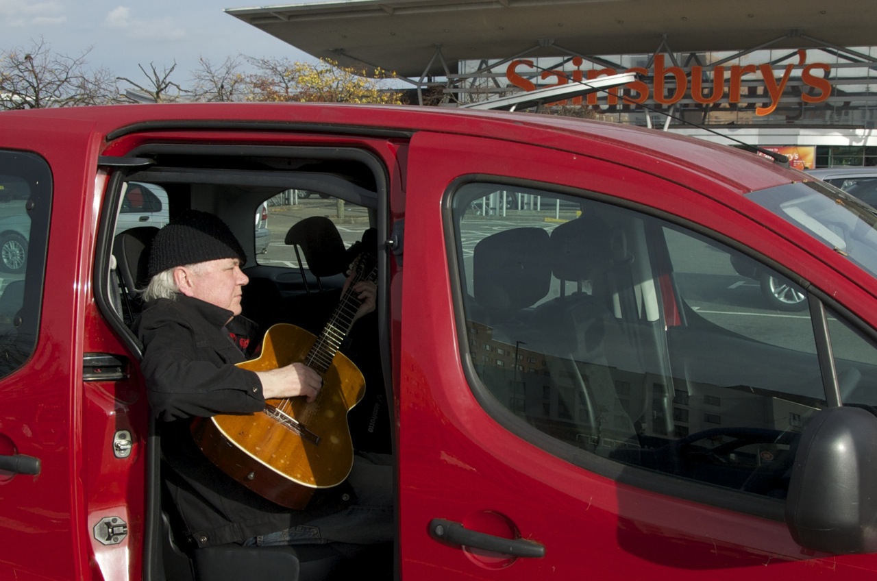 Billy Jenkins playing guitar in a supermaket car park. Photographed by Beowulf Mayfield